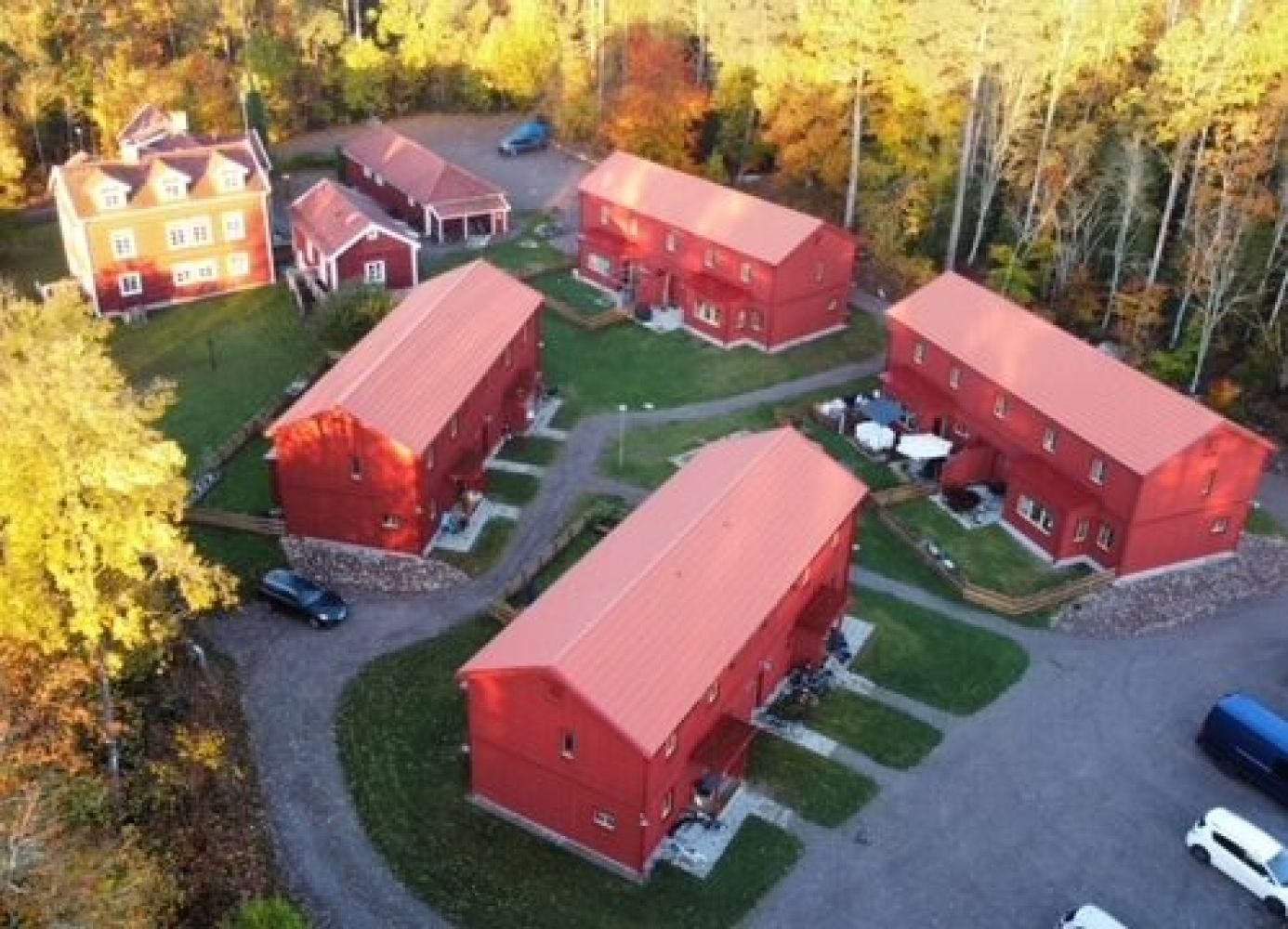 Flygfoto över bostadsområde med röda trähus i lantlig miljö, omgivna av höstfärgade träd och slingrande gångvägar mellan husen.