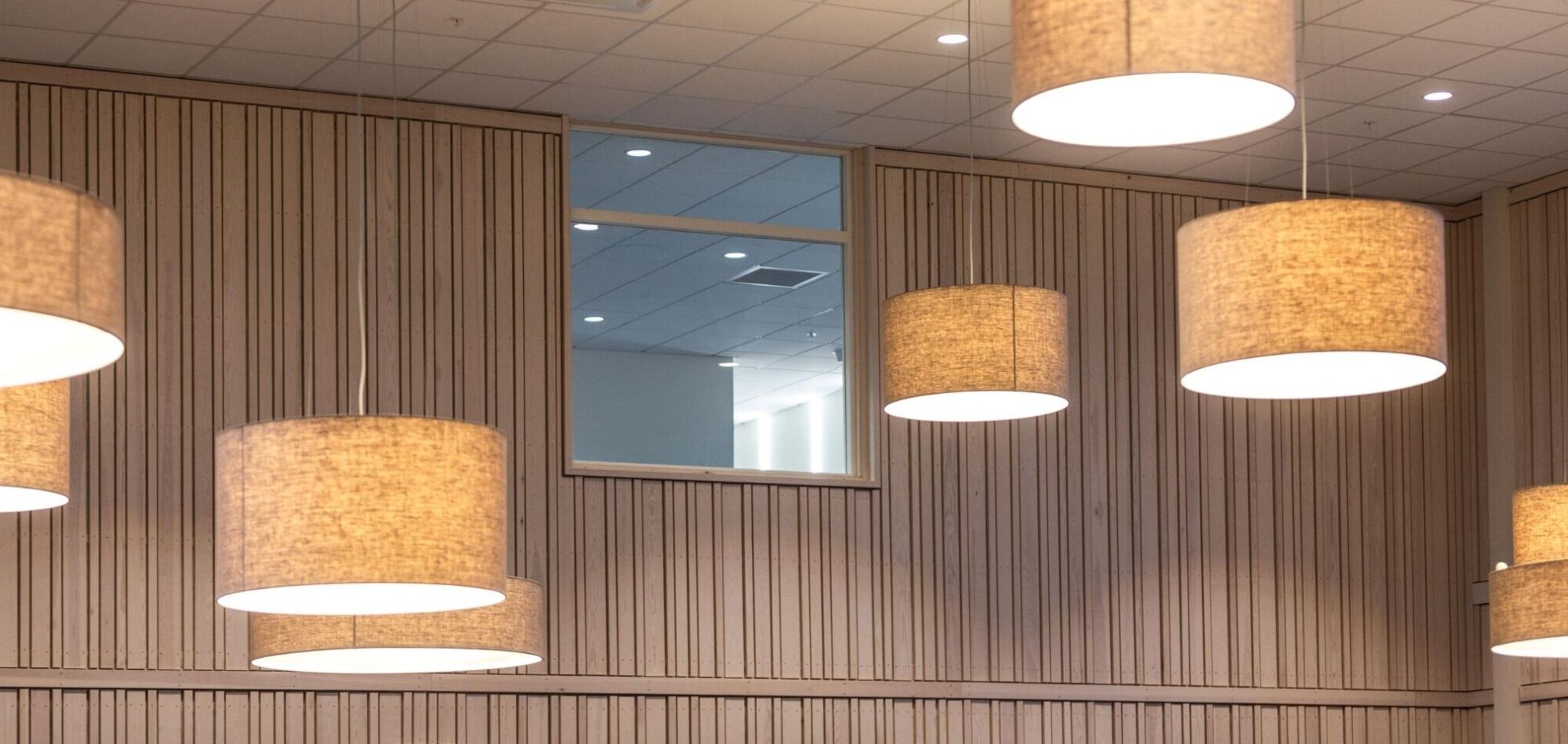 Stora pendellampor i beiget mot en träpanelvägg med fönster och taket i bakgrunden.