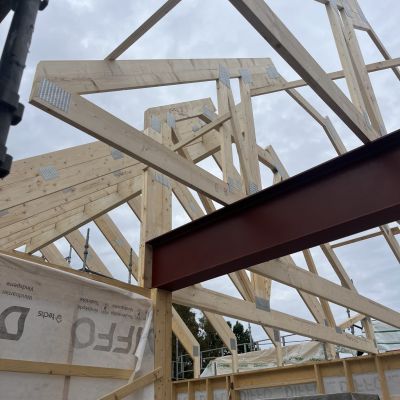 Byggarbetsplats med synlig takkonstruktion i trä och stålbalk, under uppförande mot en mulen himmel – fokus på stomme och konstruktion.