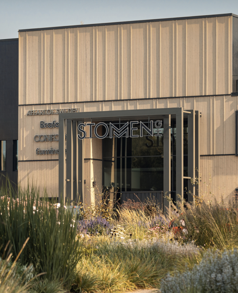Entré till byggnad med skyltningen "STOMMEN" och vertikal träpanel, omgiven av planterad grönska – modern arkitektur i harmonisk miljö.