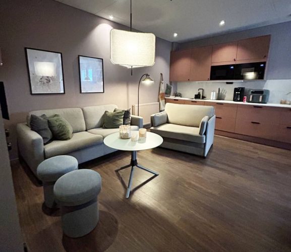 Inbjudande loungemiljö med soffa, fåtölj och runda pallar, kombinerat med ett modernt kök i dova färger och trägolv – varm och hemtrevlig inredning.