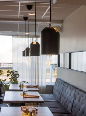 Modern matsals- eller caféinteriör med hängande svarta pendellampor, stoppad sittbänk, dukade bord och stora fönster med dagsljusinsläpp.