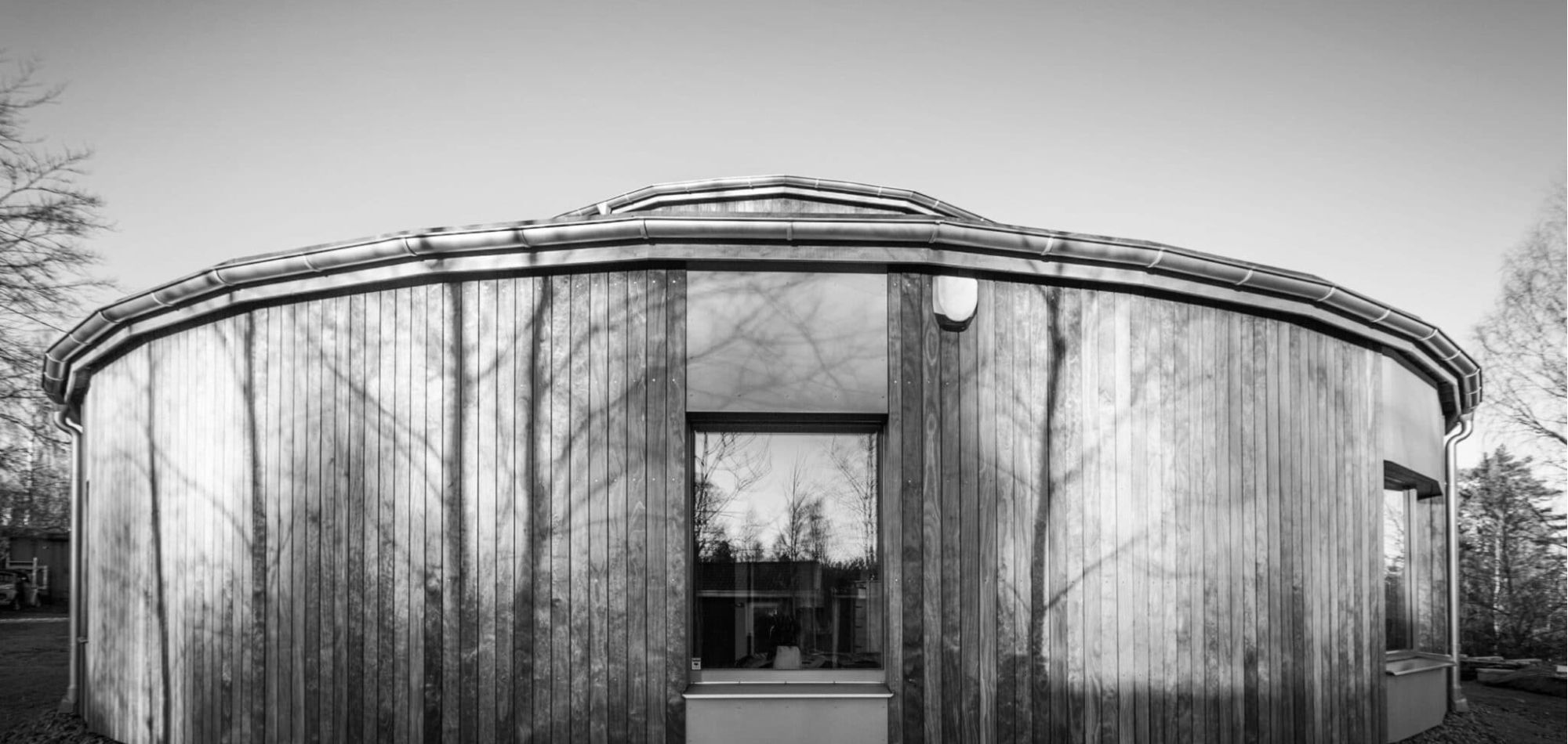 und byggnad med träfasad och stora fönster, med trädens skuggor reflekterade mot fasaden.