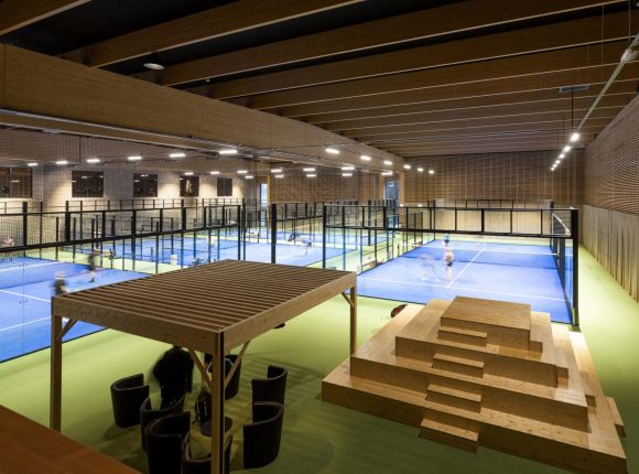 Modern padelhall med träbjälkar i taket, flera padelbanor, sittplatser och en träläktare i förgrunden – livlig aktivitet pågår på banorna.