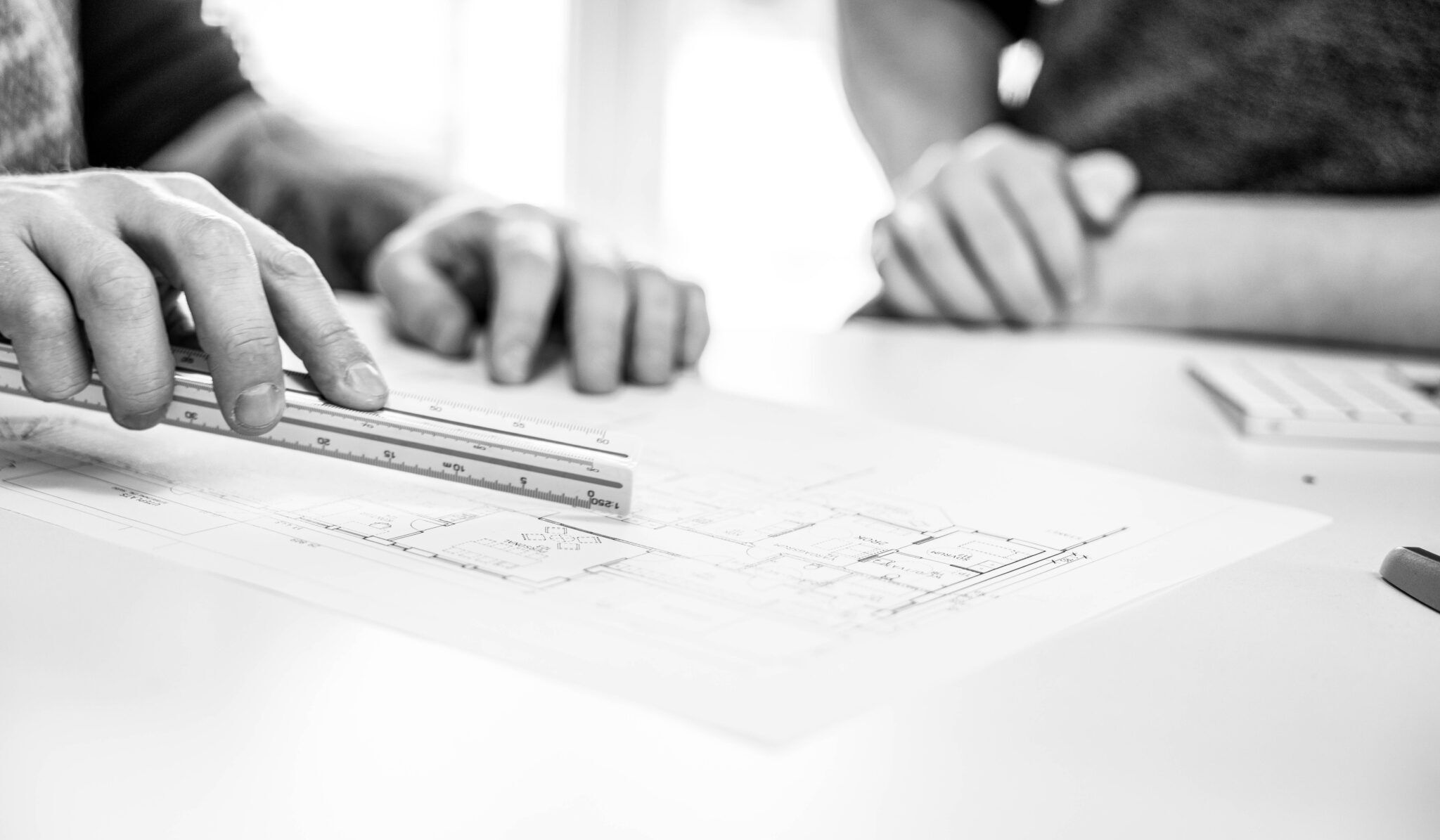 Närbild på arkitekthänder som arbetar med en ritning och en linjal på ett bord – fokus på noggrann mätning och planering.