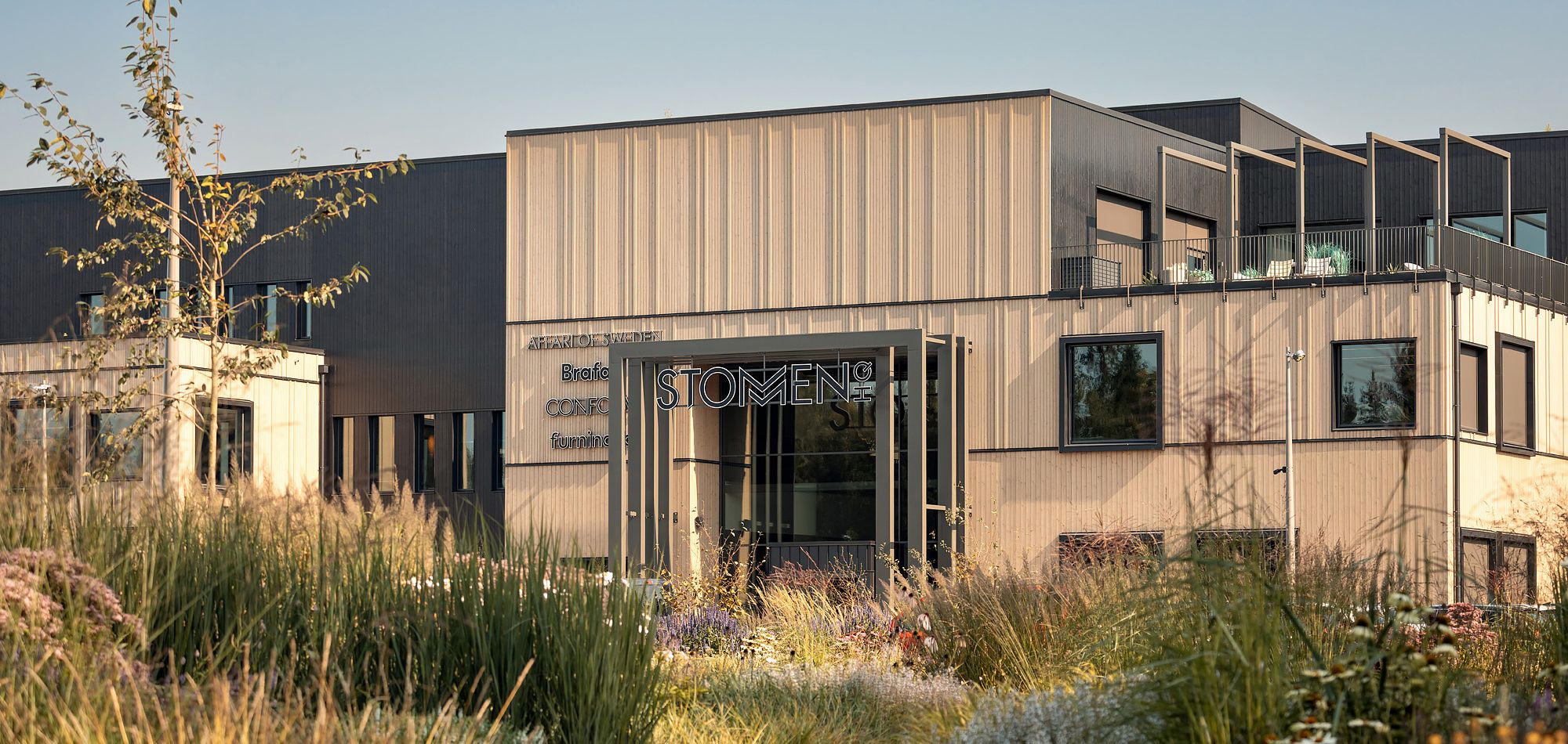 Företagshus med mörk och ljus träfasad med stor prärierabatt i förgrunden.